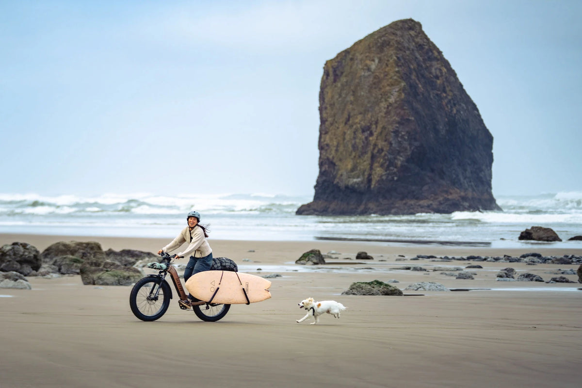 Rider carrying a surfboard on an Aventon Aventure 3 on a beach